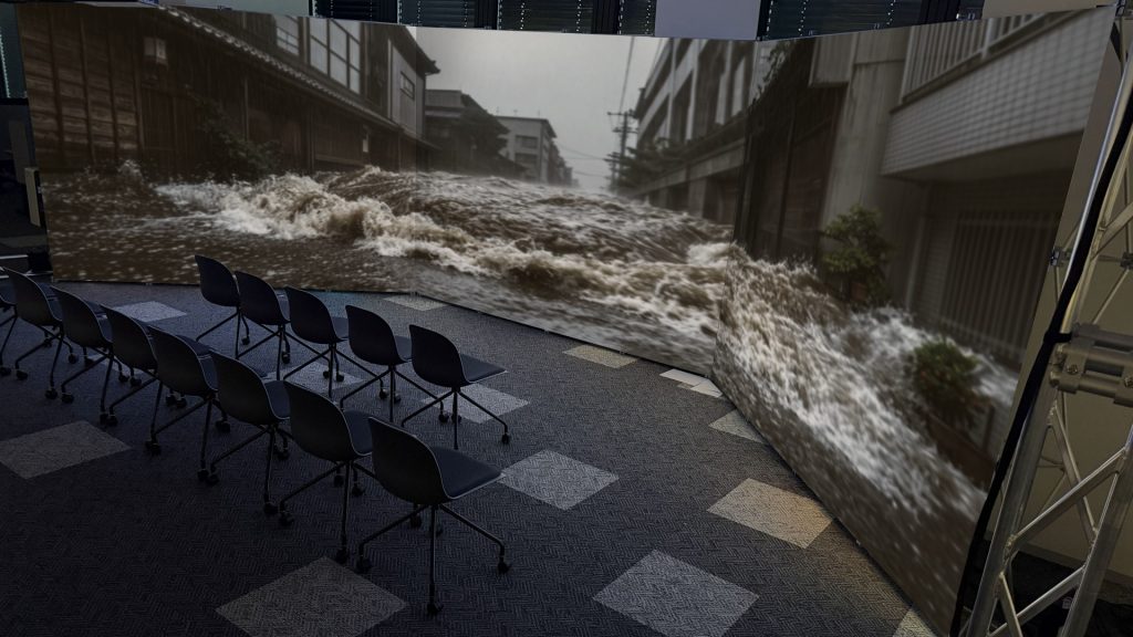 oyofair2026_1 | 株式会社キャドセンター Conference room with a curved video wall showing a flooded urban street; empty black chairs face the screen at the front of the room.
